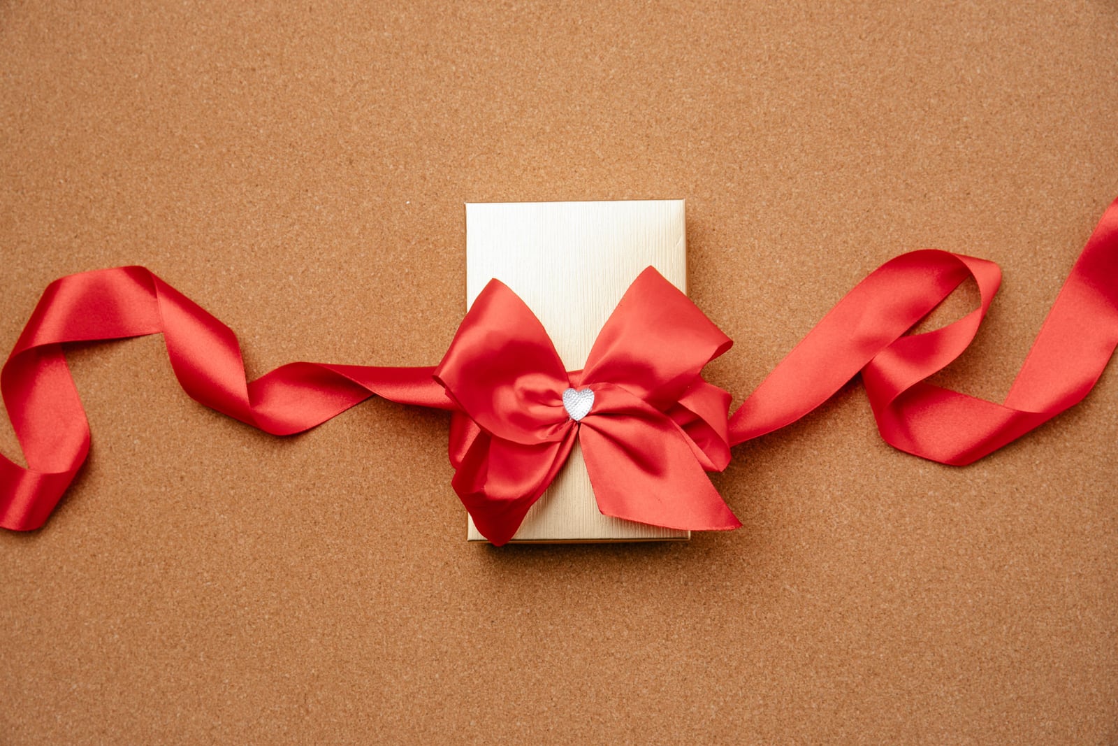 A cream-white ridged gift box with a wide red satin bow and a small silver-colored heart decoration on a textured brown surface.