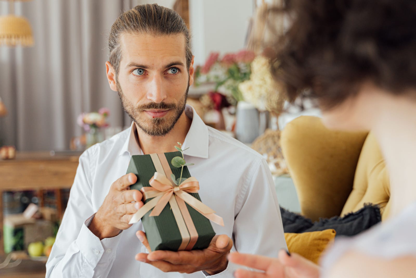 A bearded man in a white shirt offers a dark green gift box with a peach ribbon to a person seen from behind in a cozy living room.