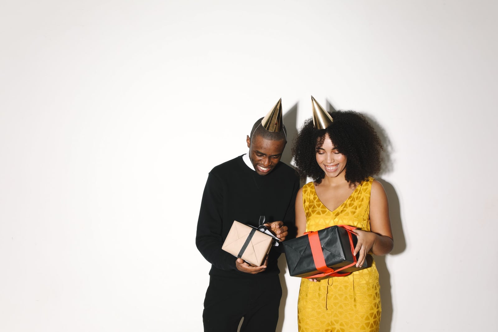 Two smiling adults in gold party hats stand together holding wrapped presents against a white wall.