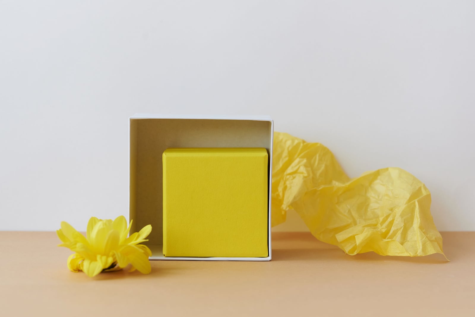 An open white box containing a yellow rectangular box on a light tabletop, with yellow tissue paper behind it and a yellow flower in front.