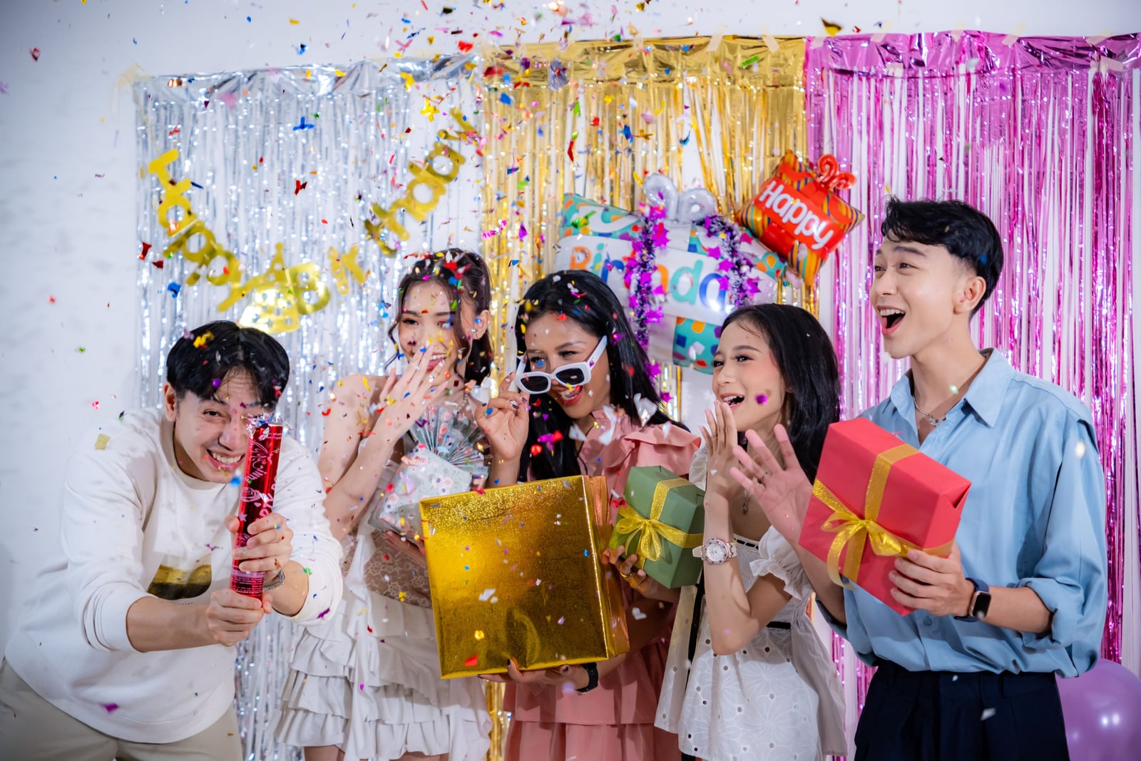 Five people pose in front of pink, silver, and gold metallic streamers as multicolored confetti falls. Some smile; one wears white sunglasses, and several hold wrapped red, green, and gold boxes.