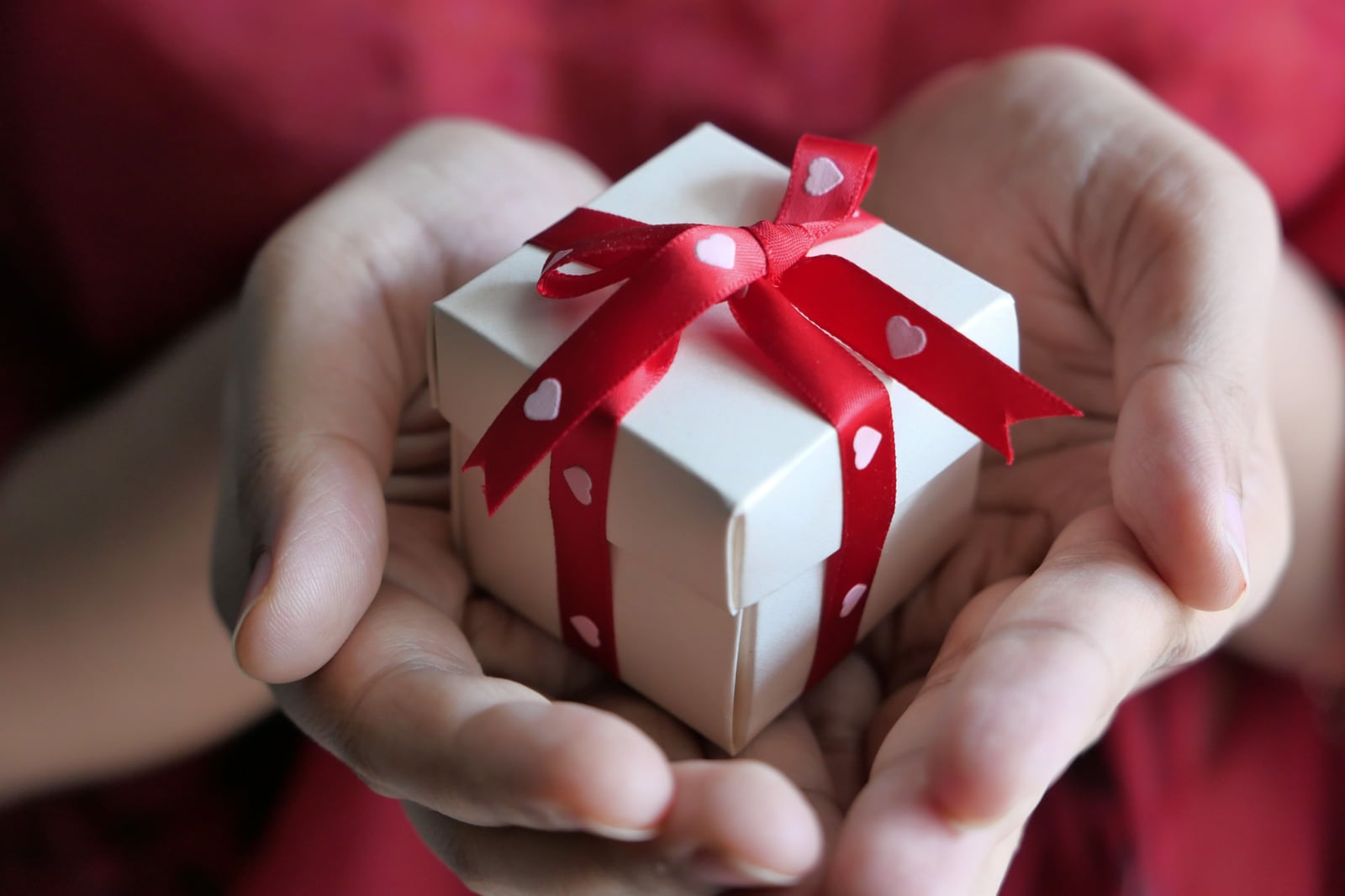 Close-up of cupped hands holding a small white square box tied with a red ribbon and bow printed with white hearts.