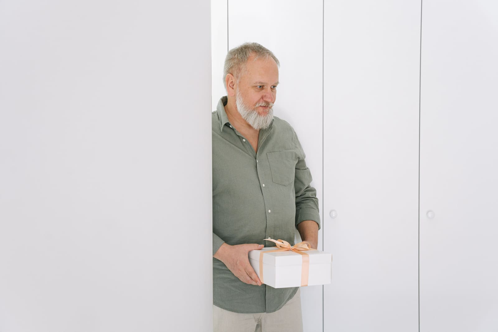 A gray-haired man peeks from behind a white door while holding a white gift box tied with a peach ribbon.