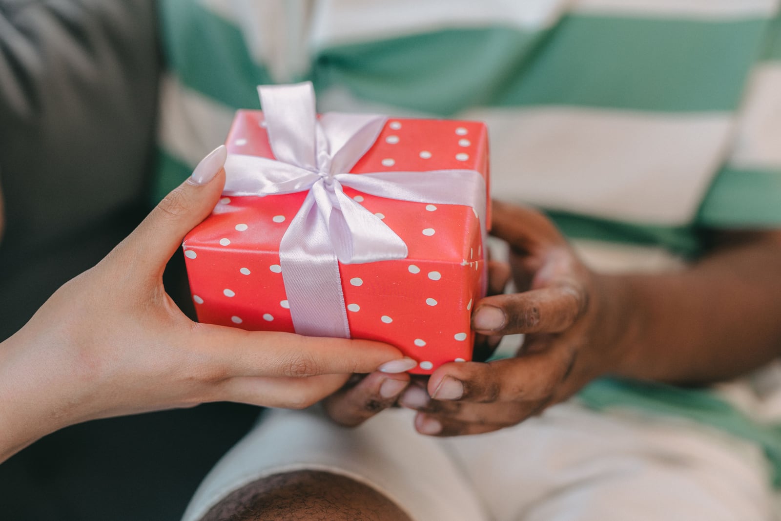 Two pairs of hands hold a red gift box with white polka dots and a pale lavender ribbon bow.