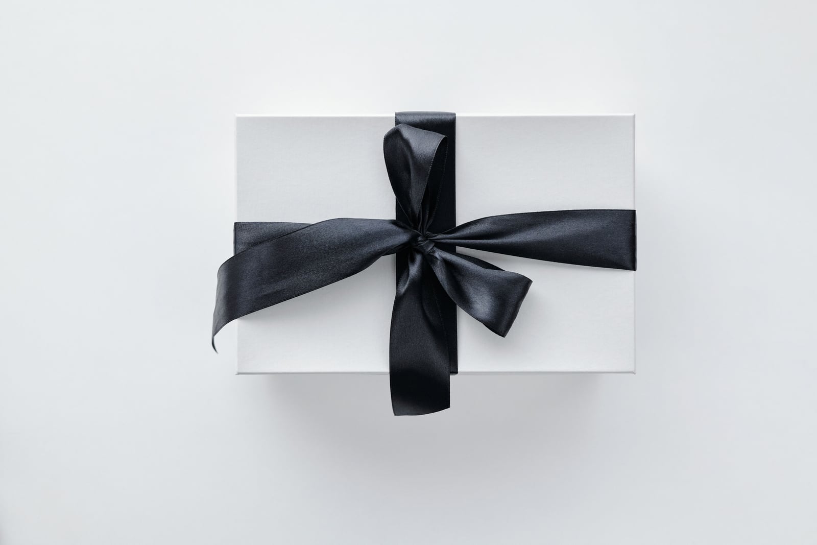 Top-down view of a white rectangular gift box wrapped with a black satin ribbon tied in a bow on a pale gray background.