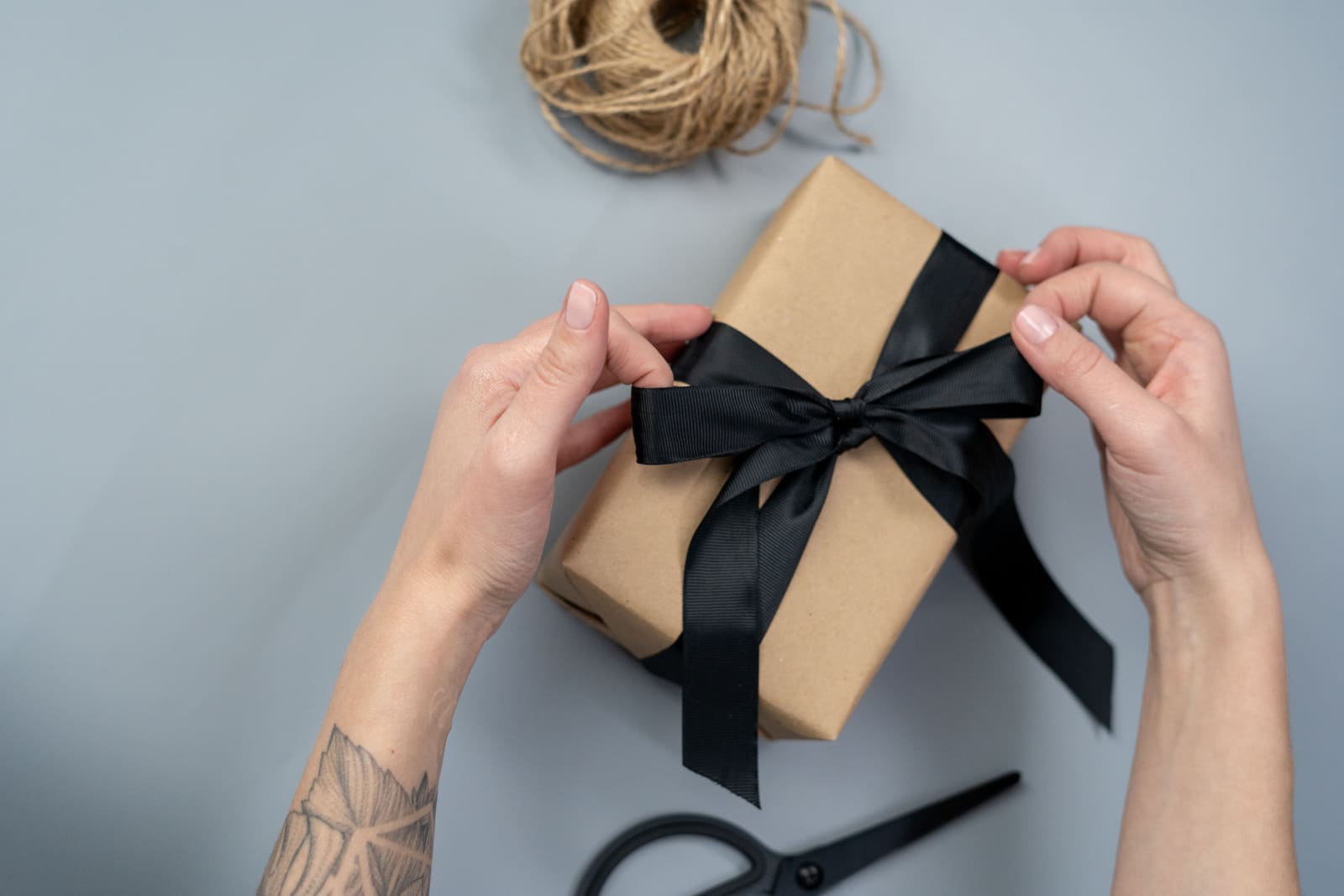 Several hands tying a dark ribbon around a kraft-paper-wrapped gift, with twine and scissors nearby.